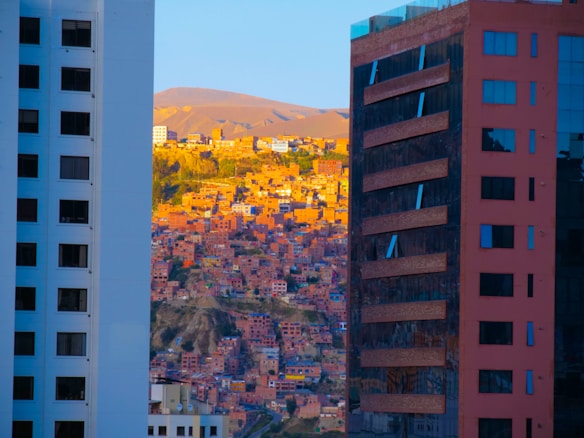 A densely built residential area with numerous small houses is nestled between two modern skyscrapers. The buildings in the foreground are tall structures with reflective surfaces, while the background features a hillside bathed in warm, golden light, creating a striking contrast between urban architecture and traditional housing.