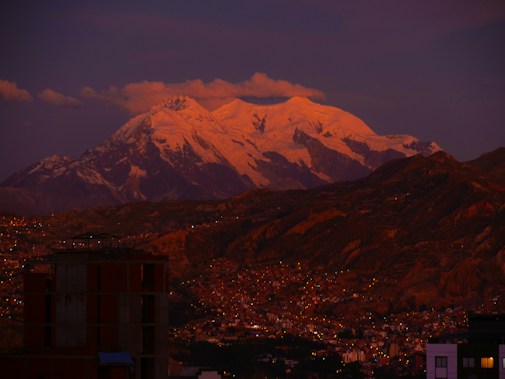 A majestic snow-capped mountain dominates the skyline, illuminated by a warm, pinkish glow from the setting or rising sun. The foreground features an expansive view of a city with buildings and twinkling lights under a darkening sky.