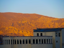 A landscape featuring a building in the foreground with large windows and a flat roof. In the background, an expansive view of a mountain range illuminated by the warm light of sunset, casting a golden glow over the rugged terrain.
