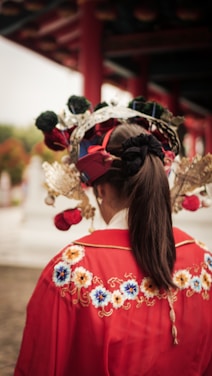 A person dressed in traditional attire featuring vibrant red fabric with intricate floral embroidery. The individual is wearing an elaborate headpiece adorned with flowers and metallic elements, and their hair is tied back in a ponytail.