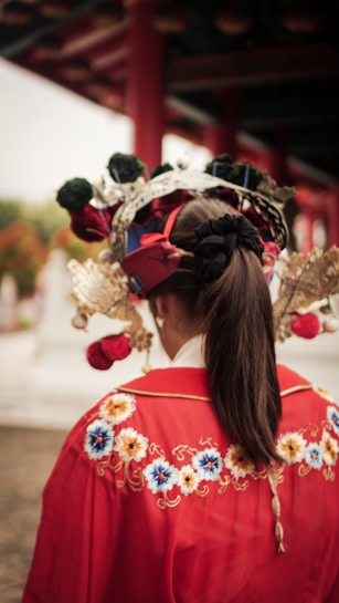 A person dressed in traditional attire featuring vibrant red fabric with intricate floral embroidery. The individual is wearing an elaborate headpiece adorned with flowers and metallic elements, and their hair is tied back in a ponytail.