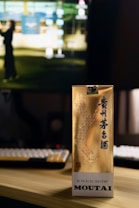 A gold-colored box with Chinese characters and an illustrated design, standing on a wooden surface in front of a computer monitor. The background is slightly blurred, showing a keyboard and part of a computer screen with a blurred image of a person.
