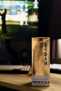 A gold-colored box with Chinese characters and an illustrated design, standing on a wooden surface in front of a computer monitor. The background is slightly blurred, showing a keyboard and part of a computer screen with a blurred image of a person.