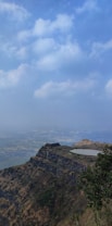 A vast landscape view featuring a ridge with an ancient fort on top and a small lake adjacent to it. The scene is set under a blue sky filled with fluffy clouds. The foreground is dominated by rugged terrain with sparse vegetation, including patches of trees.