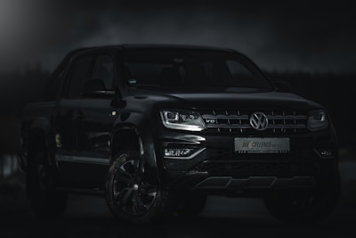 A sleek black Volkswagen pickup truck with a V6 engine badge is parked under low lighting conditions. The vehicle is positioned at an angle, highlighting its modern design and robust build. The headlights are subtly lit, and the background is almost entirely dark, giving the truck a commanding presence.