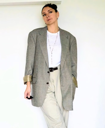 A person stands against a plain off-white background wearing a large checkered blazer with rolled-up sleeves, a white t-shirt, and khaki pants. They have short, dark hair pulled back, and they are accessorized with a long necklace and earrings. Their expression appears calm and relaxed.