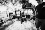 A black and white image featuring a professional camera setup mounted on a gimbal, prominently displaying the brand 'RONIN'. The scene appears to be outdoors in a wooded area with trees and dappled sunlight creating patterns on the ground. Two people are present, one partially visible holding the camera rig, wearing a cap and a t-shirt with a logo, while another person stands in the background near equipment cases.
