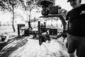 A black and white image featuring a professional camera setup mounted on a gimbal, prominently displaying the brand 'RONIN'. The scene appears to be outdoors in a wooded area with trees and dappled sunlight creating patterns on the ground. Two people are present, one partially visible holding the camera rig, wearing a cap and a t-shirt with a logo, while another person stands in the background near equipment cases.