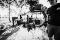 A black and white image featuring a professional camera setup mounted on a gimbal, prominently displaying the brand 'RONIN'. The scene appears to be outdoors in a wooded area with trees and dappled sunlight creating patterns on the ground. Two people are present, one partially visible holding the camera rig, wearing a cap and a t-shirt with a logo, while another person stands in the background near equipment cases.
