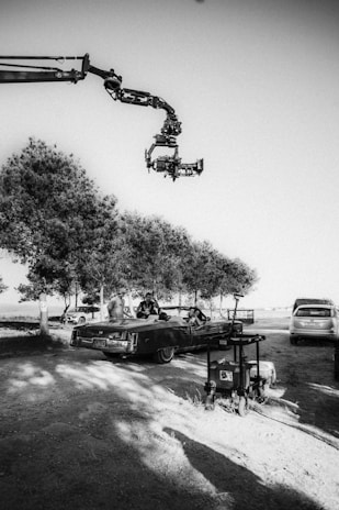 A film set featuring a large camera mounted on a crane arm is capturing scenes with a convertible car. Several individuals are gathered around the vehicle, which is parked under a row of trees. Equipment and other vehicles are also visible nearby.