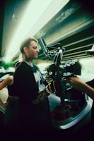A person with tattoos is operating a camera rig under a bridge. Another person is holding the equipment, and there is a car in the background with its trunk open. The setting appears to be urban, with the underside of a highway visible above.