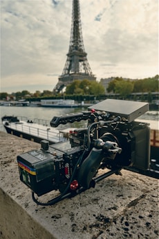 A professional video camera is positioned on a stone ledge with the Eiffel Tower and the Seine River in the background. The camera appears to be of high quality, featuring multiple lenses and attachments, and is set up for filming or photography. The scene is set against an overcast sky, providing a dramatic backdrop.