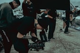 A group of people on a film set, with one individual operating a camera. They are dressed in casual clothing and appear focused. There are vehicles and equipment in the background, adding an industrial feel to the scene.