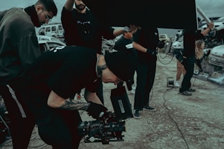 A group of people on a film set, with one individual operating a camera. They are dressed in casual clothing and appear focused. There are vehicles and equipment in the background, adding an industrial feel to the scene.