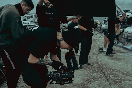 A group of people on a film set, with one individual operating a camera. They are dressed in casual clothing and appear focused. There are vehicles and equipment in the background, adding an industrial feel to the scene.