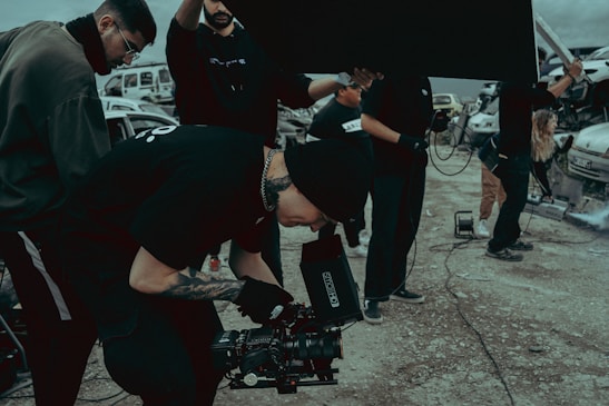 A group of people on a film set, with one individual operating a camera. They are dressed in casual clothing and appear focused. There are vehicles and equipment in the background, adding an industrial feel to the scene.