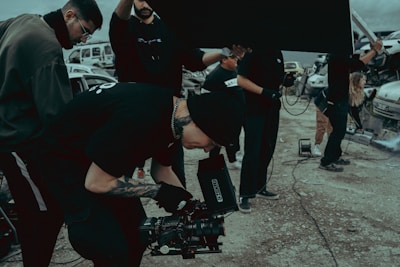 A group of people on a film set, with one individual operating a camera. They are dressed in casual clothing and appear focused. There are vehicles and equipment in the background, adding an industrial feel to the scene.