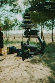 A professional film camera mounted on a mechanical stabilizing gimbal, positioned in an outdoor setting with trees and grass in the background. A person stands to the side, partially out of frame, holding a yellow object.