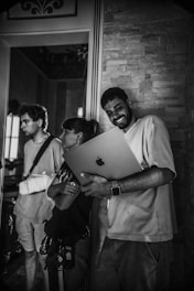 A group of three people stands indoors. One person is holding a laptop and smiling, while the others look on. The setting appears to be informal, with a stone wall and decorative elements in the background.