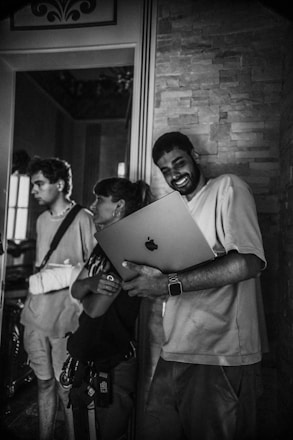 A group of three people stands indoors. One person is holding a laptop and smiling, while the others look on. The setting appears to be informal, with a stone wall and decorative elements in the background.