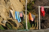 A rustic outdoor scene featuring wetsuits, towels, and clothing hanging on a clothesline strung between two wooden posts on a sandy surface. The background consists of a rocky, earth-toned cliff with some greenery and dry shrubs.