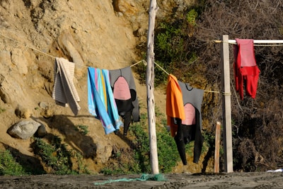 A rustic outdoor scene featuring wetsuits, towels, and clothing hanging on a clothesline strung between two wooden posts on a sandy surface. The background consists of a rocky, earth-toned cliff with some greenery and dry shrubs.