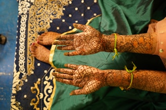 Two hands and feet are decorated with intricate henna designs, resting on a richly patterned blue and gold fabric. The person's hands are adorned with colorful wristbands, and they are wearing a green outfit with gold accents.