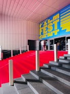 A grand staircase is covered with a bright red carpet, leading up to a building entrance. The wall above features a large, vibrant blue and yellow sign that reads 'Film Fest Gent'. The setting appears to be indoor, with a modern architectural design characterized by clean lines and a sleek ceiling. People are visible at the top of the stairs, slightly blurred, giving a sense of movement.