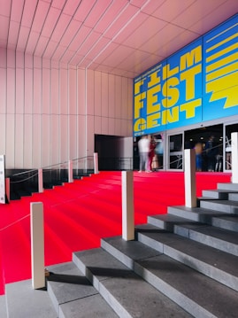 A grand staircase is covered with a bright red carpet, leading up to a building entrance. The wall above features a large, vibrant blue and yellow sign that reads 'Film Fest Gent'. The setting appears to be indoor, with a modern architectural design characterized by clean lines and a sleek ceiling. People are visible at the top of the stairs, slightly blurred, giving a sense of movement.