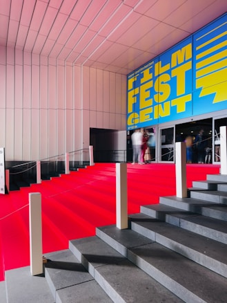 A grand staircase is covered with a bright red carpet, leading up to a building entrance. The wall above features a large, vibrant blue and yellow sign that reads 'Film Fest Gent'. The setting appears to be indoor, with a modern architectural design characterized by clean lines and a sleek ceiling. People are visible at the top of the stairs, slightly blurred, giving a sense of movement.