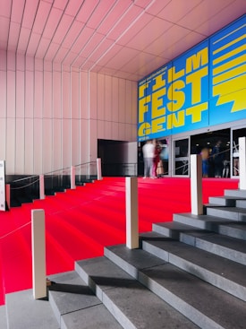 A grand staircase is covered with a bright red carpet, leading up to a building entrance. The wall above features a large, vibrant blue and yellow sign that reads 'Film Fest Gent'. The setting appears to be indoor, with a modern architectural design characterized by clean lines and a sleek ceiling. People are visible at the top of the stairs, slightly blurred, giving a sense of movement.