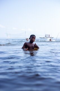 A child is partially submerged in water, holding a yellow object. The scene is set on a calm body of water with two other people visible in the background, one swimming and another near a small boat.
