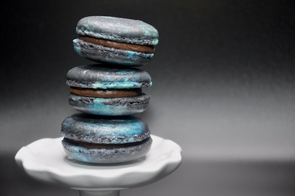 A tower of three blue and black macarons is neatly stacked on an elegant white pedestal. The macarons have a slightly rustic texture, with a creamy filling visible between their layers. The dark gradient background contrasts with the vibrant blues and subtle touches of brown in the macarons.