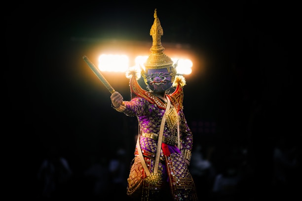 A performer in an elaborate, traditional costume stands against a dark background, illuminated by bright lights from behind. The outfit is ornate, featuring gold, purple, and red tones, with intricate patterns and a headdress.