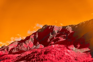 A striking landscape with rugged mountains colored in surreal hues of red and gray, set against a vibrant, orange sky. The mountains exhibit defined textures, creating a dramatic contrast with the sky. There are scattered clouds adding depth to the scene.