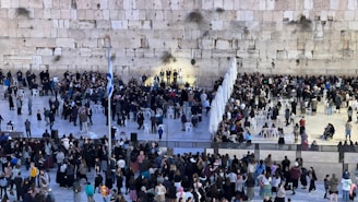A large crowd gathers in front of an ancient stone wall, with separate groups of people forming distinct sections. Many individuals are engaged in prayer or conversation, and some chairs are placed in rows. A few flags can be seen, and the scene is busy with activity.
