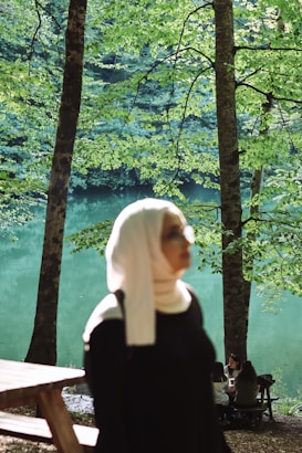 A serene lakeside setting with lush green trees framing a calm body of water. In the foreground, a person wearing a light-colored headscarf and dark clothing is slightly blurred. In the background, three people are seated at a wooden picnic table, engaged in conversation. The sunlight filters through the leaves, creating a peaceful ambiance.
