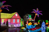 A vibrant, colorful scene featuring an illuminated red house with a yellow roof, surrounded by various tropical elements. Palm trees with vivid purple and pink leaves, surfboards with floral patterns, and a small boat add to the beach theme. Brightly lit pineapples and other decorative lights create a festive atmosphere.