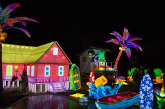 A vibrant, colorful scene featuring an illuminated red house with a yellow roof, surrounded by various tropical elements. Palm trees with vivid purple and pink leaves, surfboards with floral patterns, and a small boat add to the beach theme. Brightly lit pineapples and other decorative lights create a festive atmosphere.