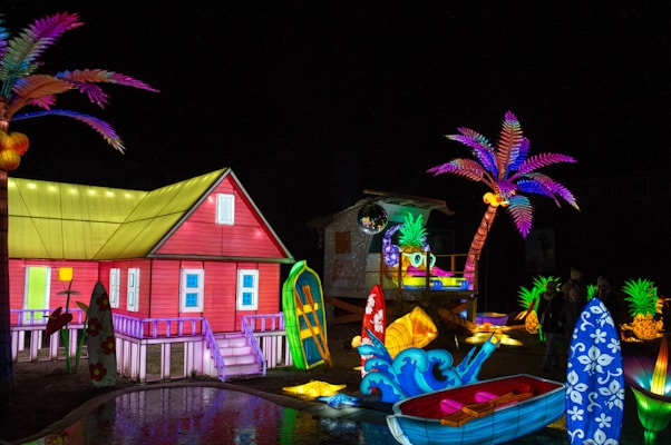 A vibrant, colorful scene featuring an illuminated red house with a yellow roof, surrounded by various tropical elements. Palm trees with vivid purple and pink leaves, surfboards with floral patterns, and a small boat add to the beach theme. Brightly lit pineapples and other decorative lights create a festive atmosphere.