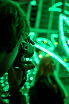 A photographer takes a picture with a camera while vibrant green lights illuminate the background. The scene focuses on a bokeh effect created by the lights, possibly of stringed or decorative type. A person stands in the background, slightly out of focus.
