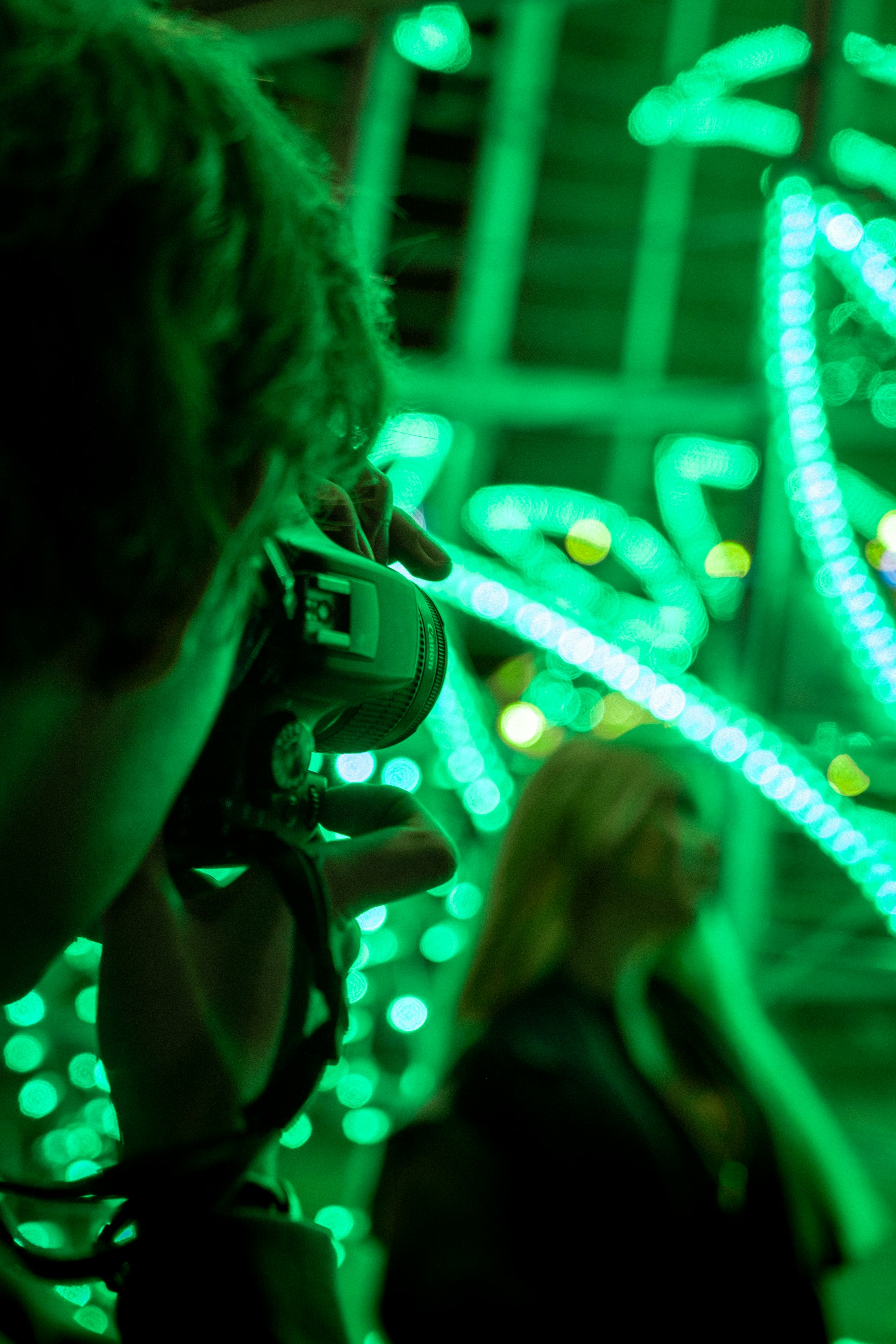 A photographer takes a picture with a camera while vibrant green lights illuminate the background. The scene focuses on a bokeh effect created by the lights, possibly of stringed or decorative type. A person stands in the background, slightly out of focus.