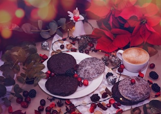 A festive arrangement featuring a plate of chocolate and powdered sugar-covered cookies surrounded by various berries, a cup of cappuccino, red poinsettia flowers, eucalyptus leaves, and small decorative ornaments. A small angel figurine holding a heart is seen in the background, adding a Christmas vibe.