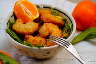 A bowl filled with crispy golden-brown chicken nuggets placed on a bed of fresh green salad leaves. An orange slice is artfully arranged next to the nuggets, and a fork rests on the bowl. Another whole orange is positioned beside the bowl on a white tablecloth.
