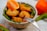 A bowl filled with crispy golden-brown chicken nuggets placed on a bed of fresh green salad leaves. An orange slice is artfully arranged next to the nuggets, and a fork rests on the bowl. Another whole orange is positioned beside the bowl on a white tablecloth.