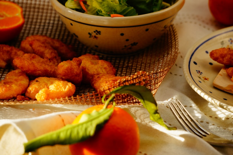 A table set up with a variety of foods, including crispy fried chicken pieces, a fresh green salad in a large bowl, and a whole ripe orange with leaves. The table is covered with a white cloth and there is a fork placed beside a ceramic plate with floral designs. The warm lighting casts soft shadows, creating a cozy and inviting atmosphere.