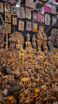 A vibrant collection of intricately crafted golden statues and religious artifacts, including various figures in meditative and symbolic poses. The display is densely packed with an assortment of sculptures, some featuring detailed engravings and colorful embellishments. On the backdrop, numerous framed artworks and decorative wall hangings complement the collection, adding texture and depth to the exhibit.