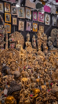 A vibrant collection of intricately crafted golden statues and religious artifacts, including various figures in meditative and symbolic poses. The display is densely packed with an assortment of sculptures, some featuring detailed engravings and colorful embellishments. On the backdrop, numerous framed artworks and decorative wall hangings complement the collection, adding texture and depth to the exhibit.