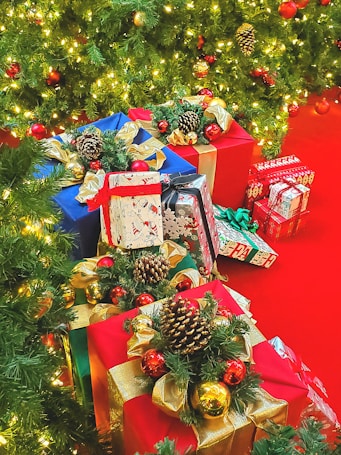 A collection of beautifully wrapped gifts is placed under a lush Christmas tree adorned with twinkling lights and red baubles. The presents are decorated with pine cones, gold ribbons, and green foliage. The floor is covered in a vibrant red carpet that complements the festive theme.