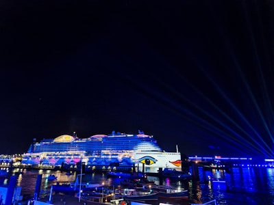 A large, illuminated cruise ship docked at a port at night, adorned with colorful lights and patterns. In the foreground, smaller boats are floating on the water. The surrounding area is also lit up with various blue and white lights, creating a vibrant and festive atmosphere.